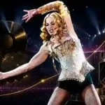 Madonna performing on stage in a sparkling silver and gold outfit, surrounded by concert lights and subtle wealth and music business imagery.